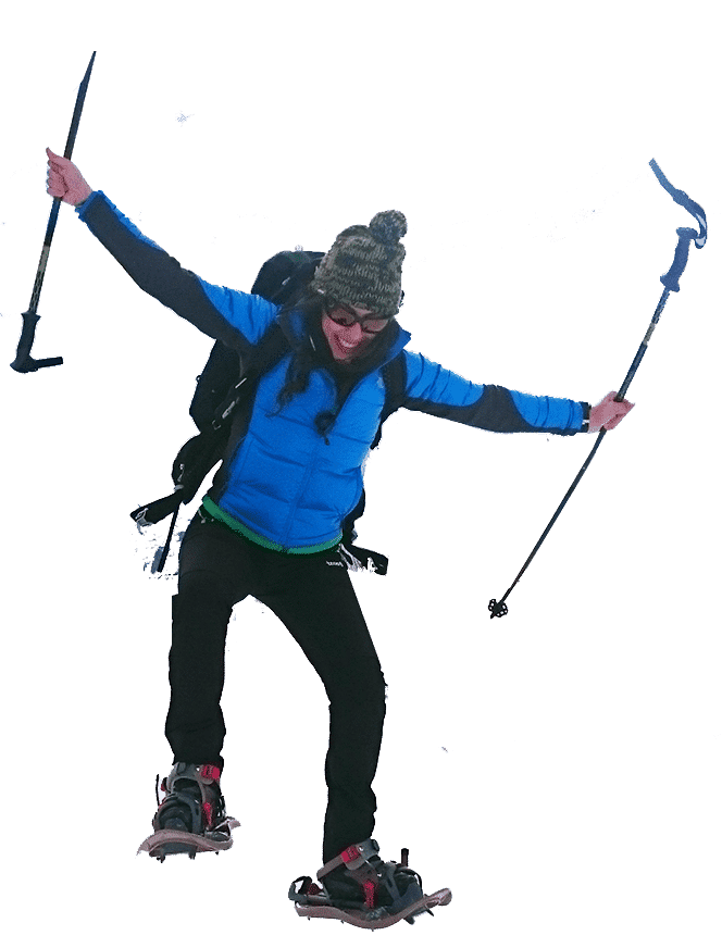 Mujer con chaqueta azul y gorro, saltando en la nieve con bastones de senderismo, mostrando entusiasmo en un entorno montañoso.